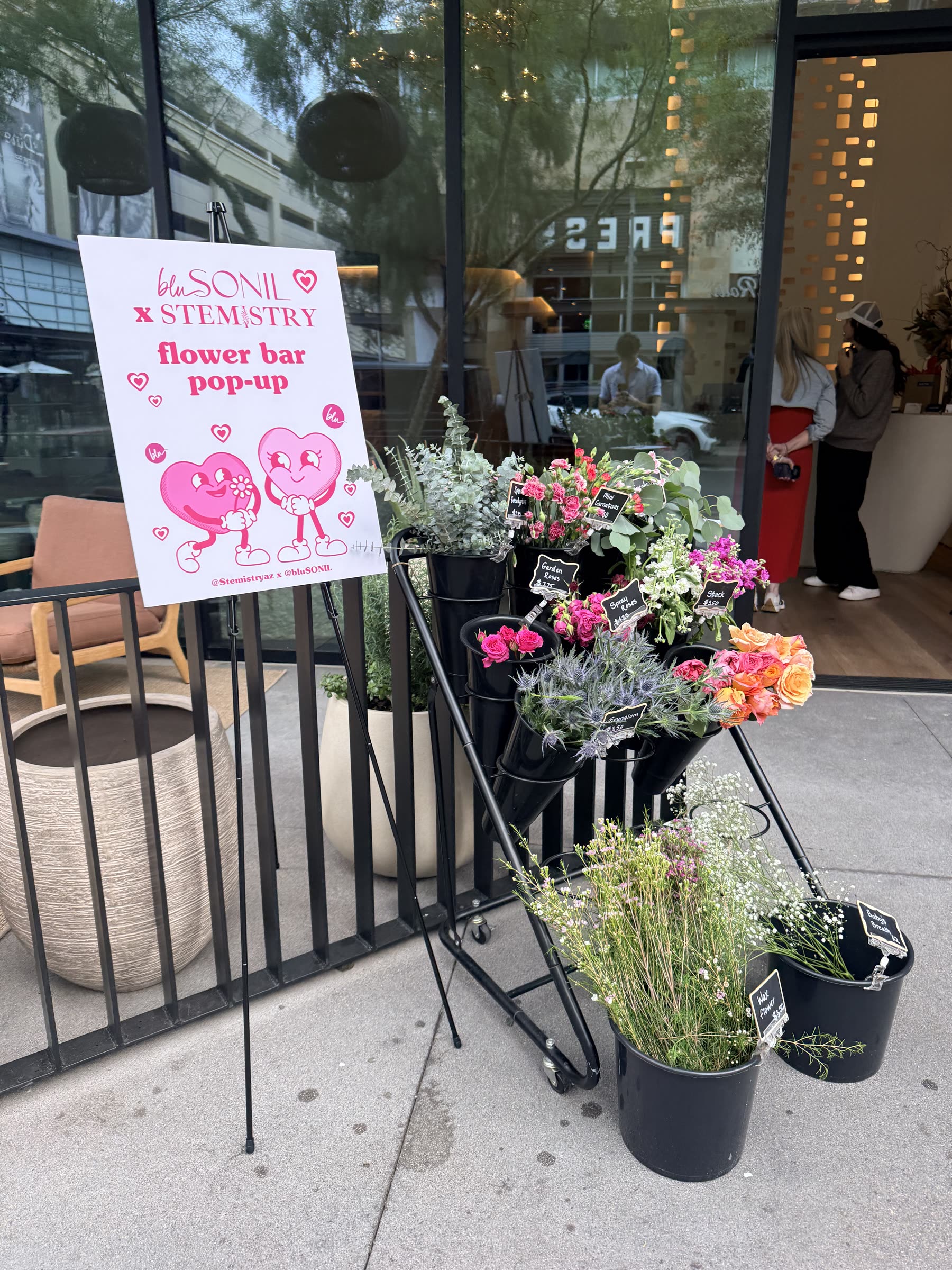 bluSONIL x Stemistry flower bar pop-up sign next to a rolling cart of roses and wax flower